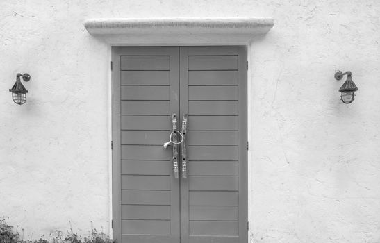 Closed wood door and white concrete wall of the building. Lamp on cement wall near the door. Building exterior. Building wall with rough texture background. Wooden door with handle at the entrance.