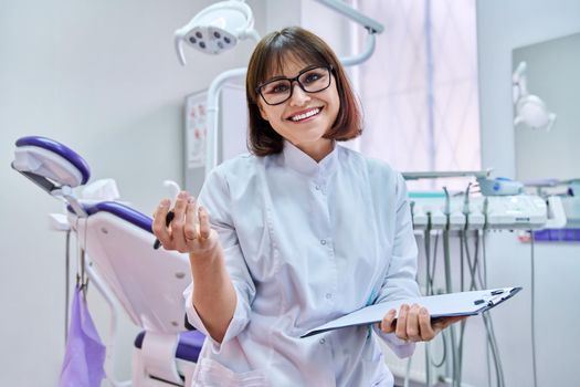 Talking consulting female doctor dentist looking at webcam, making video call chat conference, notes on clipboard, in dental office. Online technology, telemedicine, help medicine, health care concept