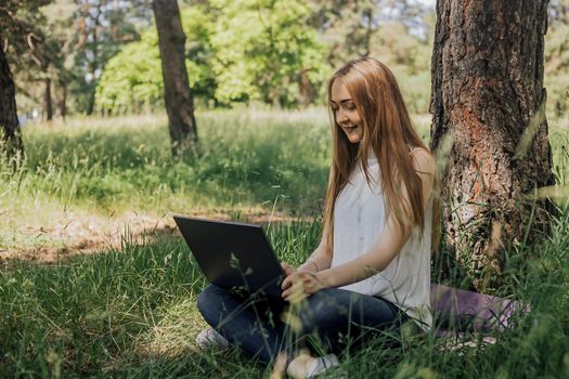 On the banner, a young girl works with a laptop in the fresh air in the park, sitting on the lawn. The concept of remote work. Work as a freelancer. The girl takes courses on a laptop and smiles