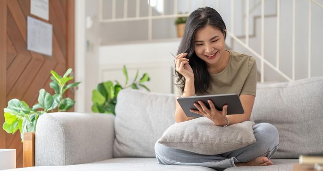 Beautiful attractive young Asian woman sitting on gray sofa and using digital tablet at living room in the morning