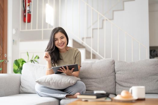 Beautiful attractive young Asian woman sitting on gray sofa and using digital tablet at living room in the morning