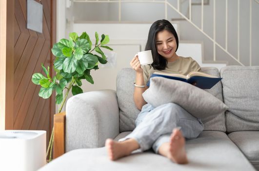 happy asian young woman sitting on sofa and reading book at home -people and leisure concept.