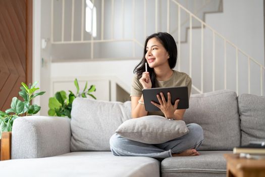 Beautiful attractive young Asian woman sitting on gray sofa and using digital tablet at living room in the morning