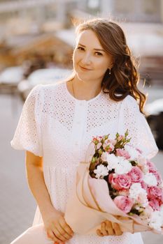 A happy woman in a white dress at sunset with a bouquet of flowers in the city.