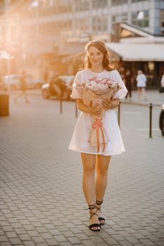 A happy woman in a white dress at sunset with a bouquet of flowers in the city.