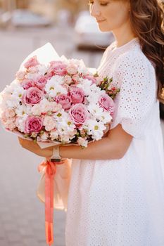 A happy woman in a white dress at sunset with a bouquet of flowers in the city.