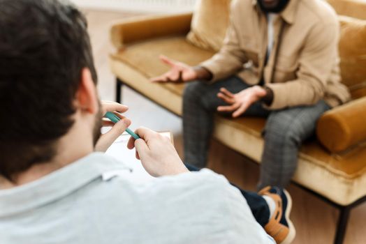 An irritated African American emotionally talks about problems to a psychologist at a session, a confused unhappy young man undergoes therapy with a psychiatrist