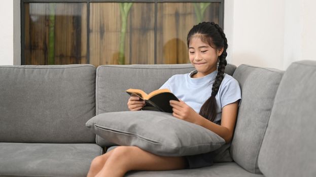 Smiling preteen girl relaxing on comfortable sofa and reading book.