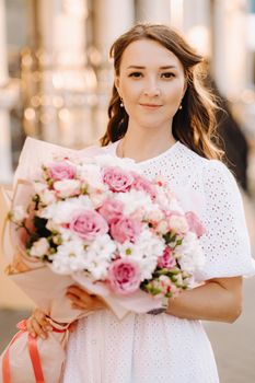 A happy woman in a white dress at sunset with a bouquet of flowers in the city.