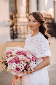 A happy woman in a white dress at sunset with a bouquet of flowers in the city.