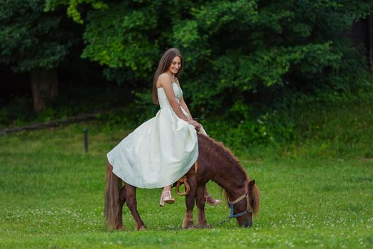 girl in a white dress rides a pony on a green lawn