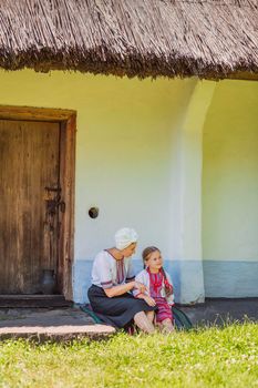 mother and daughter in Ukrainian national costumes are sitting near an old house