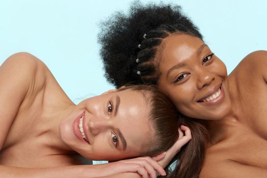 Young beautiful caucasian and african women posing lying on top of each other, face to face. Lifestyle of different nationalities, people concept. Looking at camera.