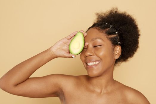 Portrait of a beautiful young African woman holding an avocado and smiling against a beige background. The concept of proper nutrition, skin care, spa and cosmetology.