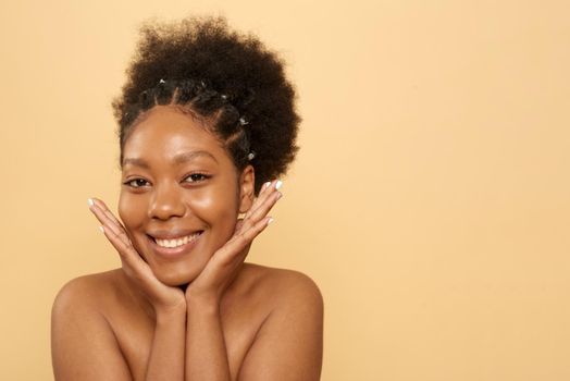 Positive young black woman with bare shoulders and beautiful skin touching face looking at camera on beige background.