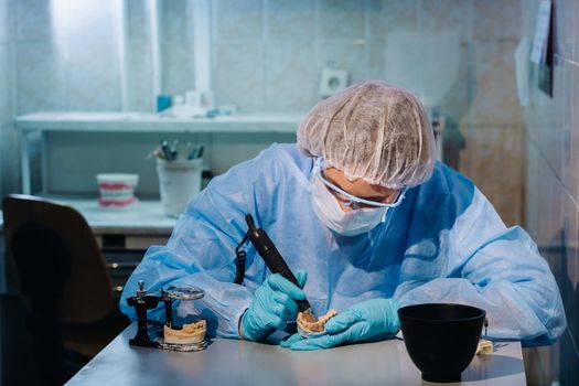 A dental technician in protective clothing is working on a prosthetic tooth in his laboratory.