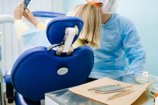 a dentist in a protective mask sits next to a patient and takes a selfie photo while working.