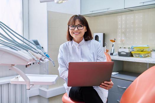 Portrait of dentist doctor sitting in office with laptop in her hands looking at camera. Mature smiling female nurse in dental clinic. Dentistry, medicine, health care, profession, stomatology concept