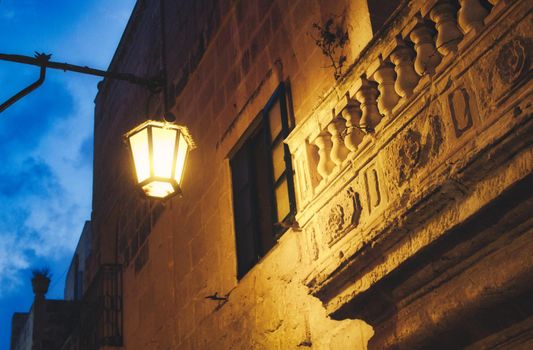 Moody shot of a yellow glowing streetlamp illuminating an old stone balcony in a dark narrow alley
