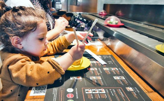 Cute little girl using chopsticks to eat sushi at a restaurant with self-service conveyor belt
