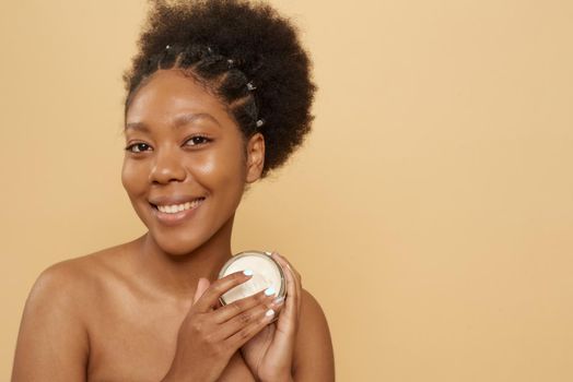 Beautiful african american woman holding jar of moisturizer or mask on beige background with copy space. Skin care, anti aging.