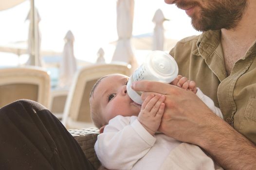 Man feeding a newborn baby with milk from a bottle at the beach on holiday