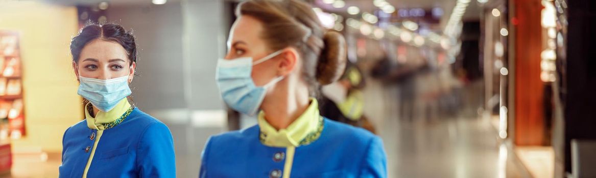 Women stewardesses wearing protective face masks and air hostess uniform while walking down airport terminal during pandemic