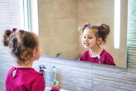 Young cute female kid looking at her reflection in the bathroom mirror while brushing her teeth