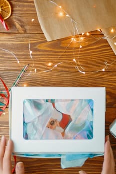 Box of Christmas gingerbread on wooden table. Women's hands reach for carton of cookies. Garland, wrapping paper and ribbons. New Year composition.