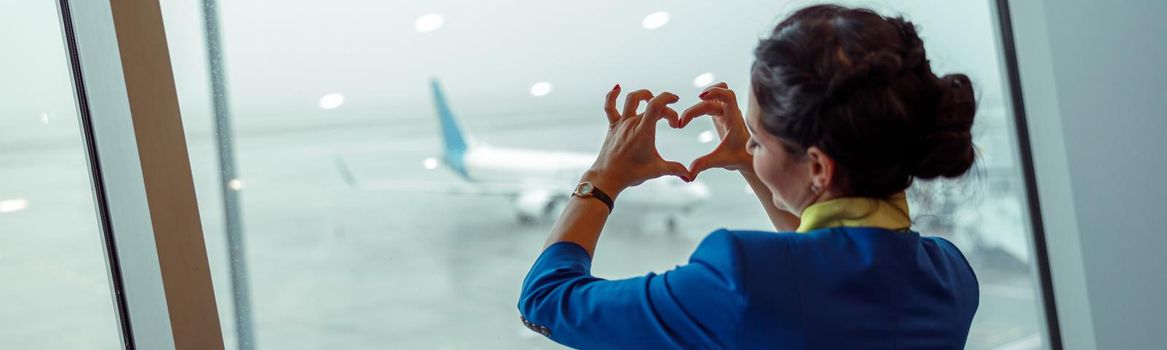 Female flight attendant making heart sign with hands while standing by the window and enjoying airfield view