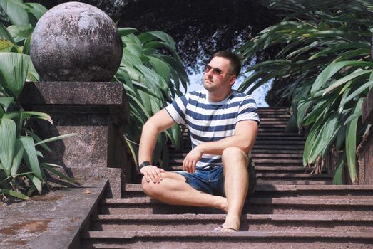 a stylish guy with glasses is sitting on the steps in a tropical garden. portrait of a young man in summer clothes. tropical country in summer. High quality photo