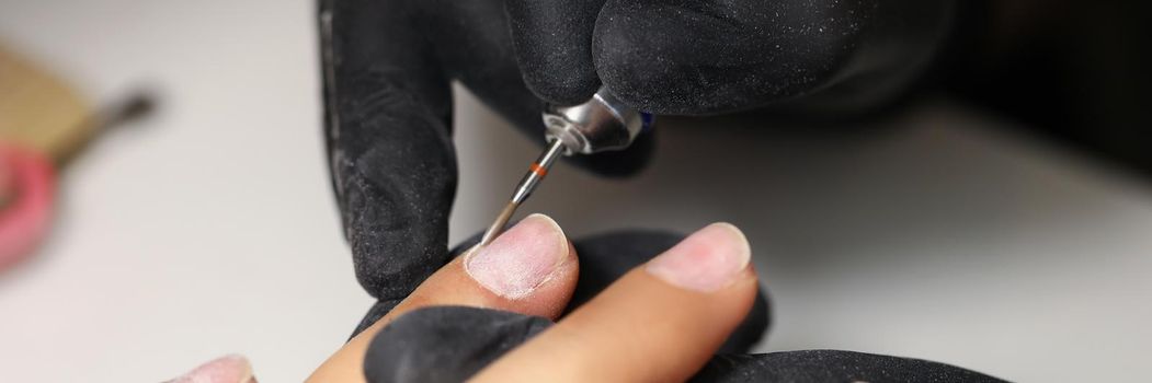 Close-up of nail master using tool to clean female clients nails in beauty studio. Worker wear protective black gloves for work. Care and beauty concept