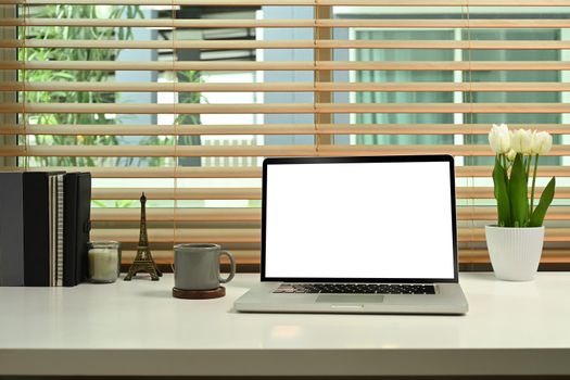 Computer laptop with white empty display, coffee cup, flower pot and books on home office desk.