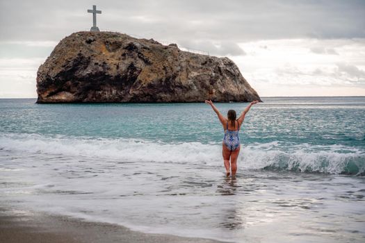 A plump woman in a bathing suit enters the water during the surf. Alone on the beach, Gray sky in the clouds, swimming in winter