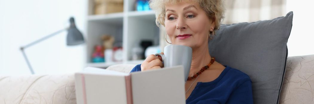 Portrait of old lady reading book and drinking cup of coffee being comfy on sofa at home. Attractive senior woman relaxing on weekend. Spare time concept