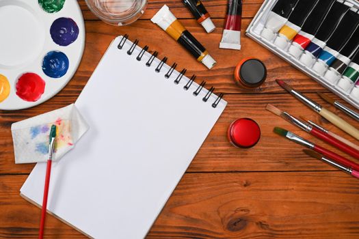 Artist workplace with paint brushes, watercolor paints, notepad and palette on wooden background. Top view.