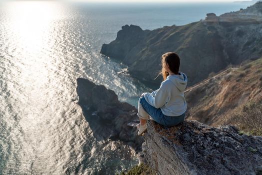 Woman tourist enjoying the sunset over the sea mountain landscape. Sits outdoors on a rock above the sea. She is wearing jeans and a blue hoodie. Healthy lifestyle, harmony and meditation.