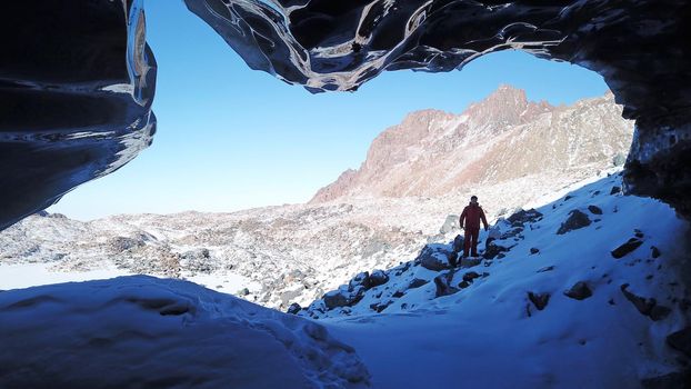 A climber looks at the entrance to an ice cave. Frozen snow mountains and glacier. Entrance looks like a dolphin. High peaks, clear blue sky and big rocks. An extreme journey. Lots of snow underfoot