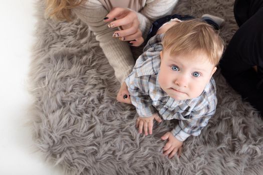 A one-year-old blond baby crawls on the carpet, surrounded by parents, looks at the camera. Top view.