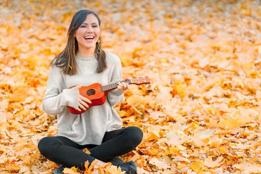 Young happy woman having a good time with musical instrument sitting on fallen autumn leaves.