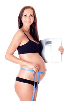 Beautiful smiling pregnant woman measuring her tummy by tape measure and holding scales. Isolated on white.