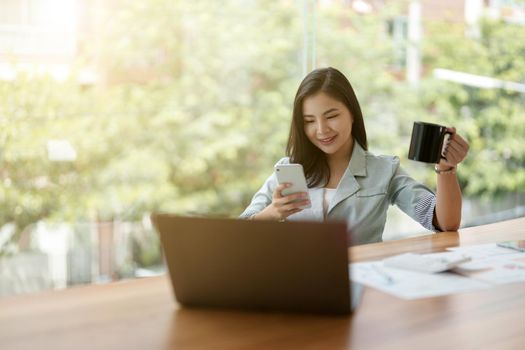 Beautiful young asian business woman using phone while working with calculator and financial paperwork in office.