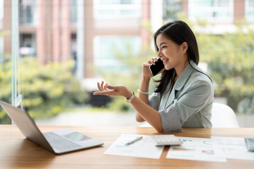 Corporate Communication. Asian Businesswoman Talking On Mobile Phone Working On Laptop In Modern Office