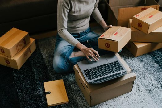 Asian female online store small business owner entrepreneur seller packing shipping ecommerce box checking website retail order using laptop preparing delivery parcel on table. Dropshipping concept