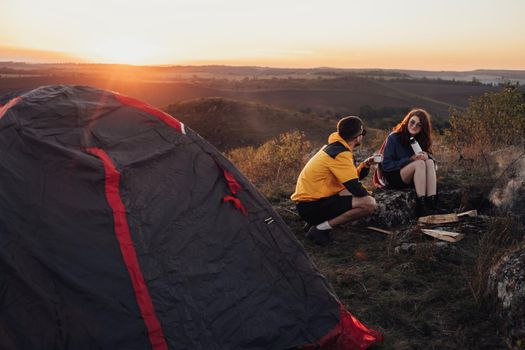 Two Young Travelers Man and Woman Sitting Near Campfire and Tent During Sunset Over Top of Hill