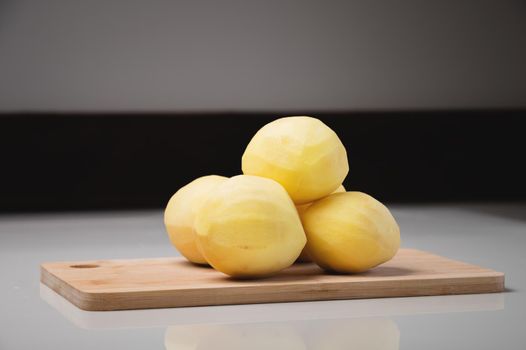 Peeled fresh potatoes lie on a wooden cutting board on a white table.