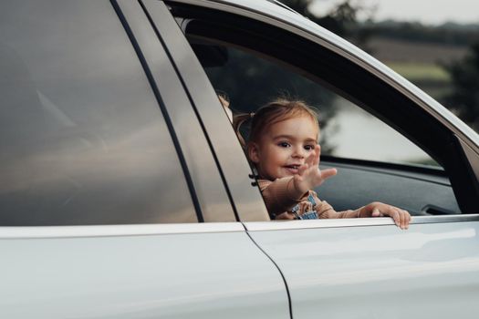 Little Girl Looking Out of Car Window and Waves Her Hand Showing Hello