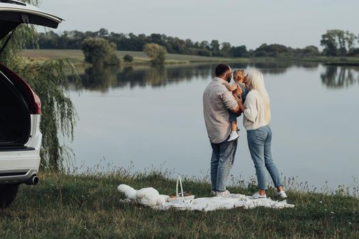 Young Parents with Their Toddler Daughter Hugging Near the SUV Car, Family Enjoying Road Trip Weekend Outside City Near the Lake