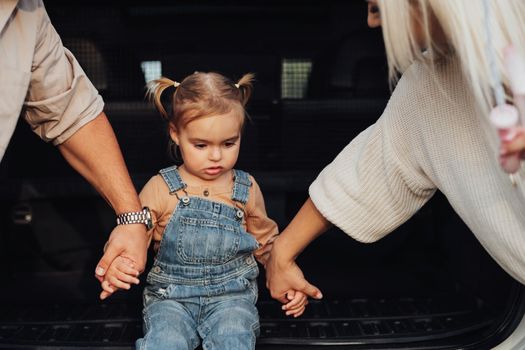 Little Girl Sitting in Open Trunk of Car, Her Parents Holding Her by Hands and Helping to Step Up