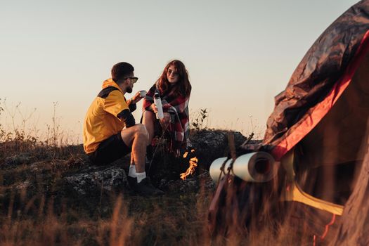 Two Young Travelers Man and Woman Sitting Near Campfire and Tent Outdoors During Sunset and Drinking Hot Tea from Thermos